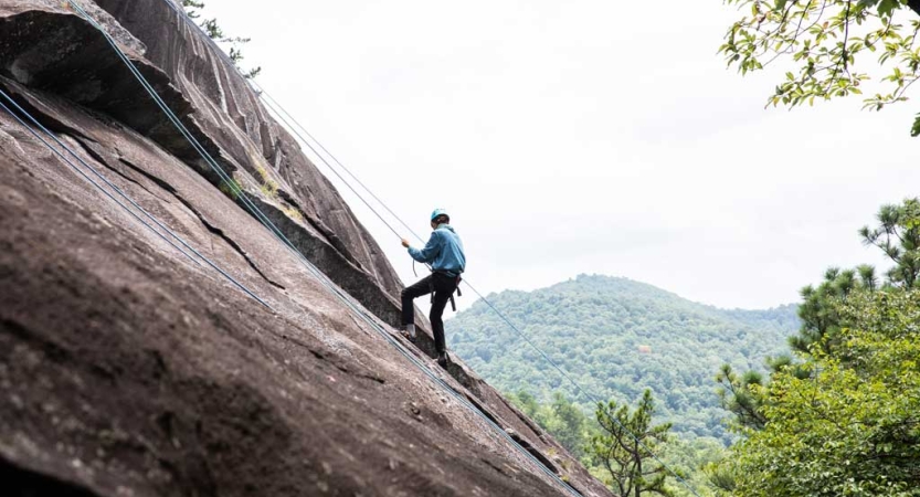 A student wearing safety gear climbs up a rock incline in front of green trees.